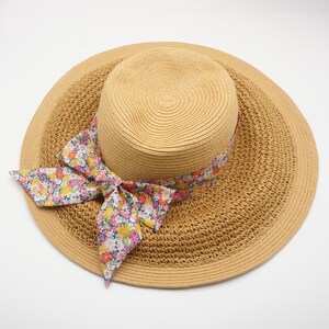 May include: A woven straw sun hat with a wide brim and a floral ribbon. The hat is a light tan color, and the ribbon features a colorful pattern of flowers in pink, yellow, and blue. The hat is designed for sun protection.