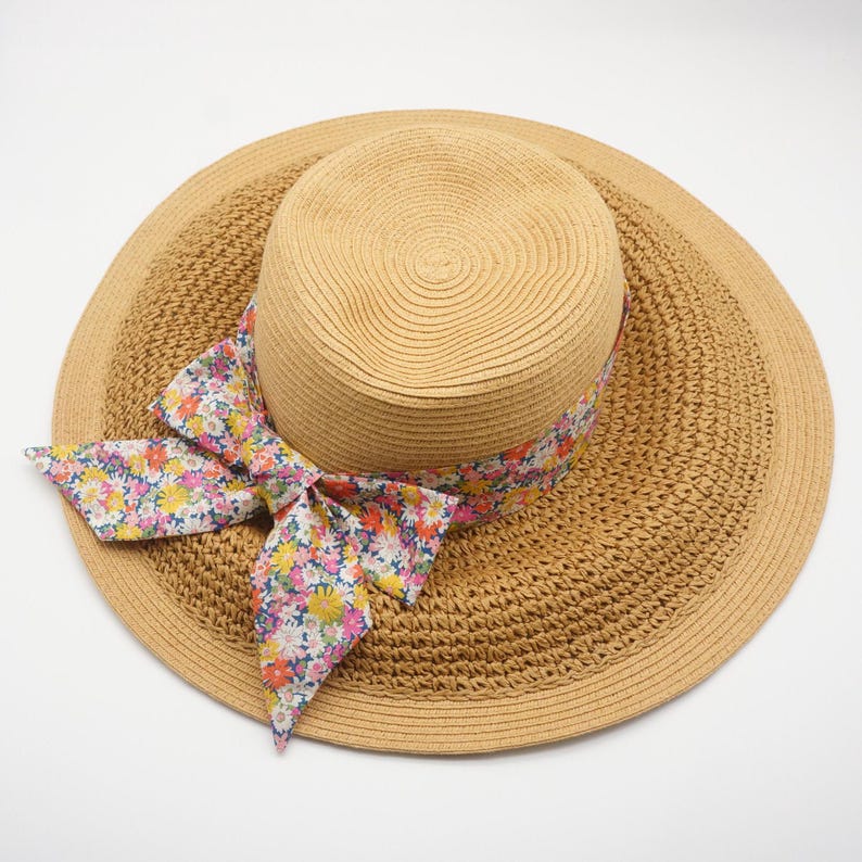 May include: A woven straw sun hat with a wide brim and a floral ribbon. The hat is a light tan color, and the ribbon features a colorful pattern of flowers in pink, yellow, and blue. The hat is designed for sun protection.