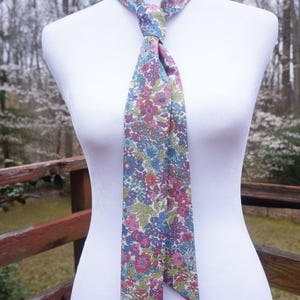 May include: A floral patterned necktie in shades of pink, blue, and green, tied in a knot. The tie is displayed on a white mannequin, with a wooden fence and trees in the background. The tie's design features a repeating pattern of flowers and leaves.