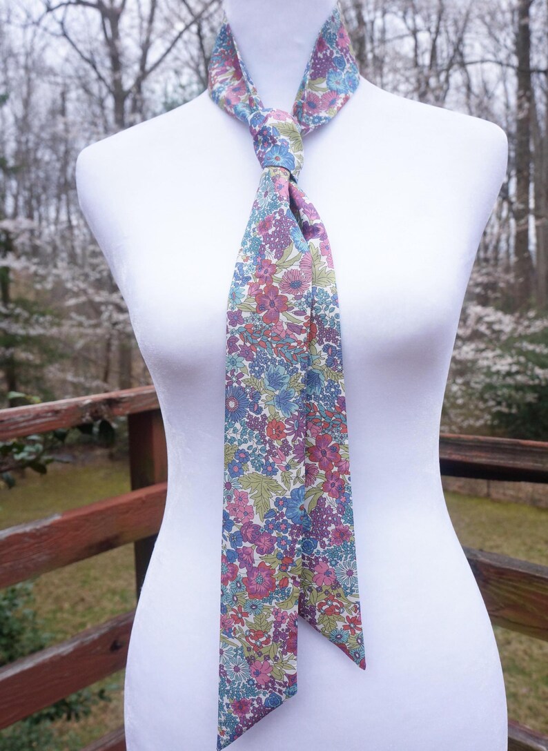 May include: A floral patterned necktie in shades of pink, blue, and green, tied in a knot. The tie is displayed on a white mannequin, with a wooden fence and trees in the background. The tie's design features a repeating pattern of flowers and leaves.
