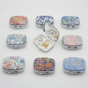May include: A collection of small, rectangular jewelry boxes with floral fabric tops and silver-colored metal frames. One box is open, revealing a mirror and compartments holding rings and necklaces. The boxes feature various floral patterns in pink, blue, and orange.