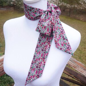 May include: A floral patterned neck scarf in shades of pink, purple, and green, tied in a bow. The scarf is displayed on a white mannequin, showcasing its design and shape. The fabric appears to be lightweight, suitable for a decorative accessory.