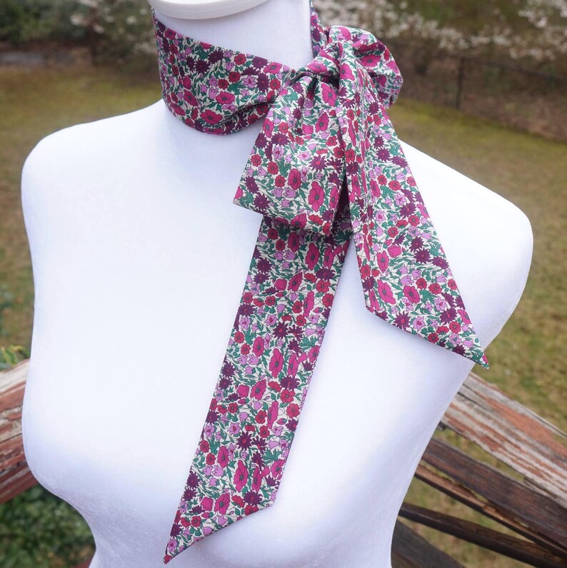 May include: A floral patterned neck scarf in shades of pink, purple, and green, tied in a bow. The scarf is displayed on a white mannequin, showcasing its design and shape. The fabric appears to be lightweight, suitable for a decorative accessory.