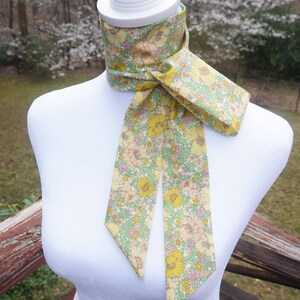May include: A floral patterned neck scarf in yellow, pink, and green, displayed on a white mannequin. The scarf features a repeating pattern of flowers and leaves. The scarf is tied in a knot, with the ends hanging down.