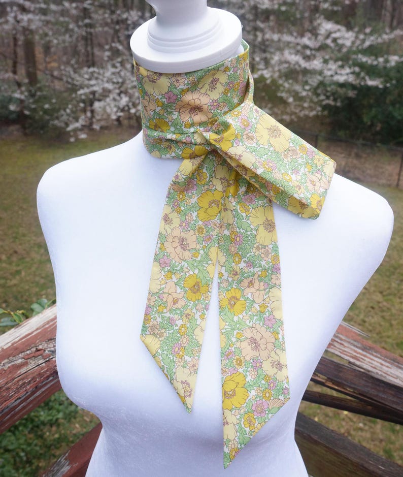 May include: A floral patterned neck scarf in yellow, pink, and green, displayed on a white mannequin. The scarf features a repeating pattern of flowers and leaves. The scarf is tied in a knot, with the ends hanging down.
