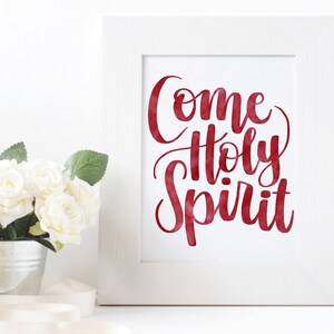 May include: White frame with a red watercolour print that says "Come Holy Spirit". The frame is sitting on a white surface with a white ribbon and a small metal bucket with white roses.