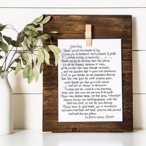 May include: A framed print with a handwritten prayer, held by a wooden clip on a dark brown wooden board. A white vase with green leaves is to the left. The text is in black script.
