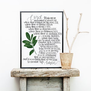 May include: A black framed print with a white background and black calligraphy text. The text reads "Lord make me an instrument of your peace...to eternal life. Amen." A green leafy branch is in the upper left corner of the print. The print is sitting on a rustic wooden shelf with a small brown pot and a branch.
