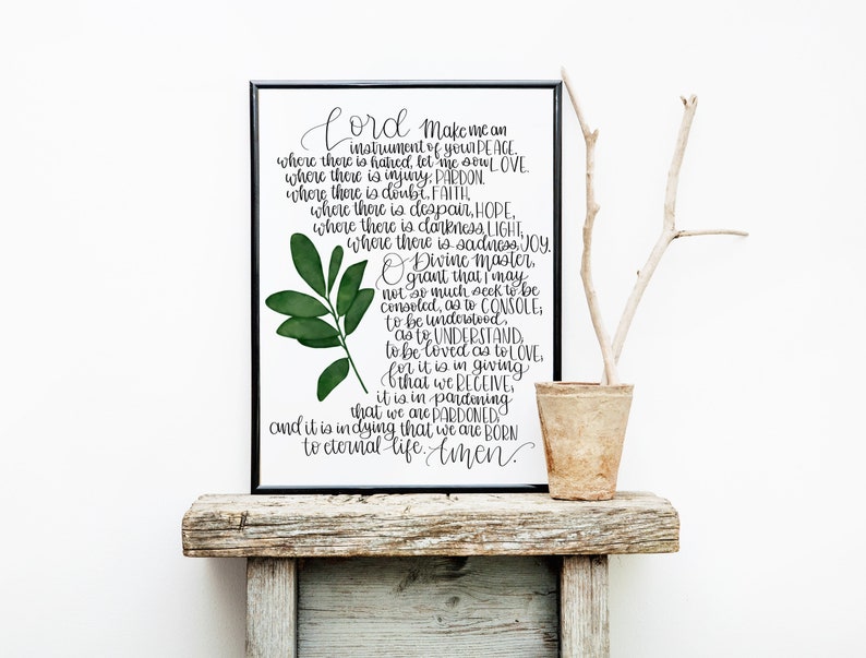 May include: A black framed print with a white background and black calligraphy text. The text reads "Lord make me an instrument of your peace...to eternal life. Amen." A green leafy branch is in the upper left corner of the print. The print is sitting on a rustic wooden shelf with a small brown pot and a branch.
