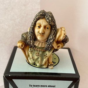 May include: A detailed figurine of King Louis XIV, with long, wavy hair and a mustache, wearing a detailed historical outfit. The figurine is on a black box with text that reads "To learn more about Pot Bellys, visit www.HarmonyBall.com".
