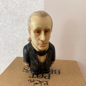 May include: A small figurine of a man with white hair and a black suit. The figurine is standing on a box with the text "Harmony Ball Co." and "BE POT" printed on it.