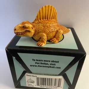 May include: A tan and brown Razor Back dinosaur figurine with a prominent dorsal fin, resting on a black box with text. The box has a light blue border and the text "To learn more about Pot Bellys, visit www.HarmonyBall.com".