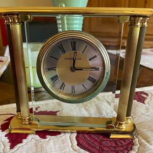 May include: A gold-toned clock with Roman numeral markings on the face. The clock is encased in a glass frame with gold pillars on either side. The clock face reads "Howard Miller".