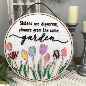 Puede incluir: Un letrero redondo de madera con fondo blanco y el texto "Sisters are different flowers from the same garden" en letras negras. El letrero está decorado con tulipanes coloridos.