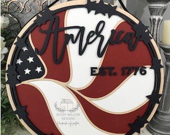 May include: A round wooden sign with a red, white, and blue American flag design. The sign has a black border with black stars around the edge. The words "America Est. 1776" are written in black on the sign.