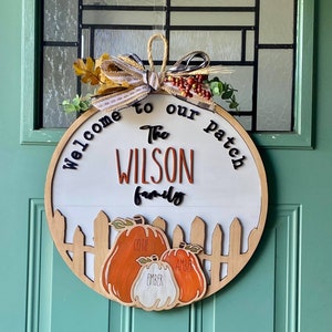 May include: Welcome to the Wilson family patch, a wooden round sign with a fall theme. The sign features a white background with black lettering and a brown wooden frame. The sign has a brown ribbon with a black and white plaid bow and fall foliage. The sign also features three pumpkins with the names "Coffe", "Ember", and "Amber" written on them.
