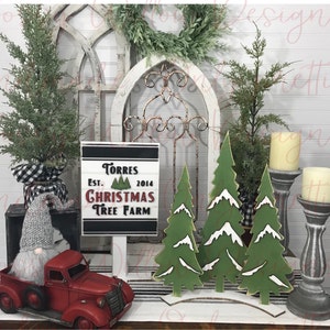 Puede incluir: Un camión de juguete rojo con un gnomo blanco en la caja. Un letrero de madera con rayas blancas y negras dice "Torres Est. 2014 Christmas Tree Farm". Tres árboles de Navidad de madera verde con acentos de nieve blanca están sobre una superficie de rayas blancas y negras. Dos candelabros grises con velas blancas están en el lado derecho de la imagen.