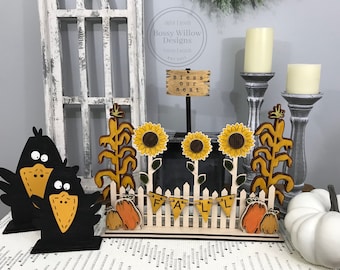 May include: A wooden fall-themed decor piece with a white picket fence, sunflowers, corn stalks, and two black crows. The fence has a banner that reads "FALL". There are also two pumpkins on either side of the fence. The sign above the fence reads "Bless our nest".