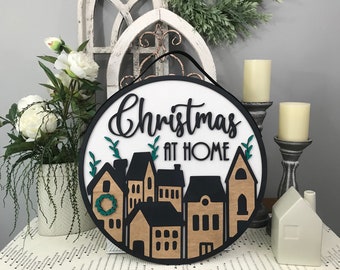 Puede incluir: Un letrero redondo de madera con fondo blanco y letras negras que dice "Navidad en casa". El letrero está decorado con una silueta negra de casas y una corona verde.