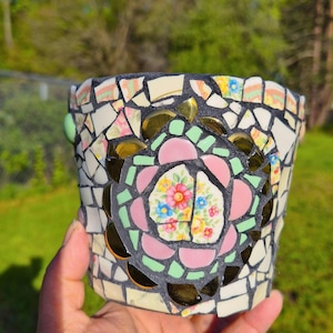 May include: A mosaic flower pot with a central floral design. The pot is made of various ceramic pieces in shades of pink, green, gold, and white. The pot's rim is black, and the overall design is intricate and colorful.