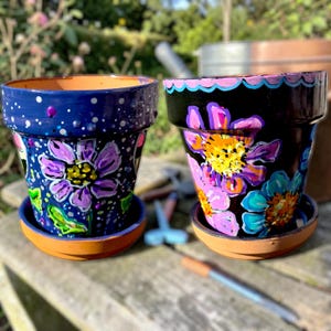 May include: Two hand-painted flower pots with matching saucers. One pot is navy blue with white polka dots and purple flowers. The other is black with colorful flowers. Both pots are on a wooden surface.