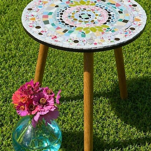 May include: A round side table with a mosaic top featuring colorful floral and geometric tiles in shades of pink, green, blue, and white, supported by three light brown wooden legs, accompanied by a clear blue glass vase filled with pink flowers.