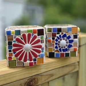 May include: Two square mosaic planters featuring colorful glass tiles. The left planter displays a large red flower design with a white center. The right planter features a blue floral pattern with a white center, surrounded by various multicolored tiles.