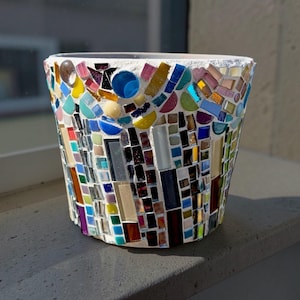 May include: A tapered cylindrical planter covered in a mosaic of multicolored glass and ceramic tiles. The exterior features a dense arrangement of rectangular, square, and circular pieces in shades of blue, yellow, green, pink, and brown set in white grout.
