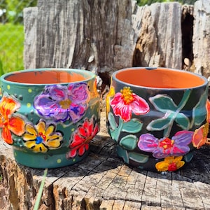 May include: Two hand-painted ceramic flower pots. One pot is teal with orange, yellow, and purple flowers. The other pot is black with pink, purple, and yellow flowers. Both pots have an orange interior.