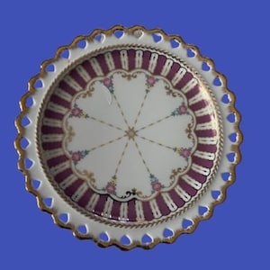 May include: A white and gold decorative plate with a scalloped edge featuring heart-shaped cutouts. The plate's center has a white background with a purple and gold design, and floral accents.