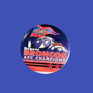 May include: A round Denver Broncos pin with a blue background. The pin features the text "1997", "Denver Broncos AFC Champions", and a graphic of a horse. The pin has red, white, and blue accents.