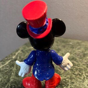 May include: A Mickey Mouse figurine viewed from the back. The figure wears a red top hat, a blue jacket, red shoes, and white gloves. The figure's ears are black. The figurine is on a green and brown surface.
