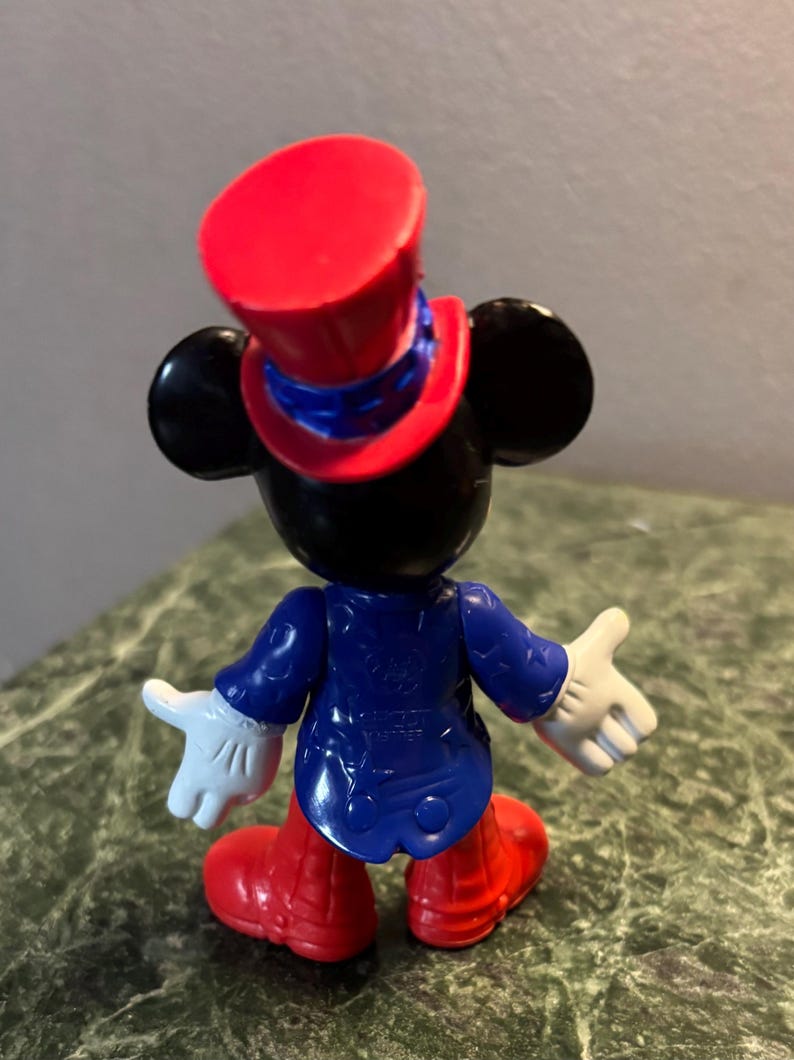 May include: A Mickey Mouse figurine viewed from the back. The figure wears a red top hat, a blue jacket, red shoes, and white gloves. The figure's ears are black. The figurine is on a green and brown surface.
