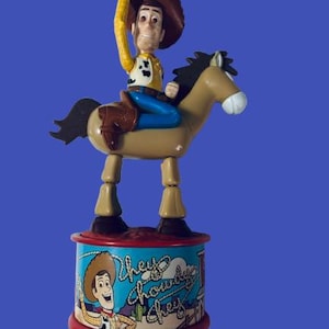May include: A Woody toy figure riding a horse, with a brown hat and yellow shirt. The base features the text "Hey Howdy Hey" and an image of Woody. The toy is set against a blue background.