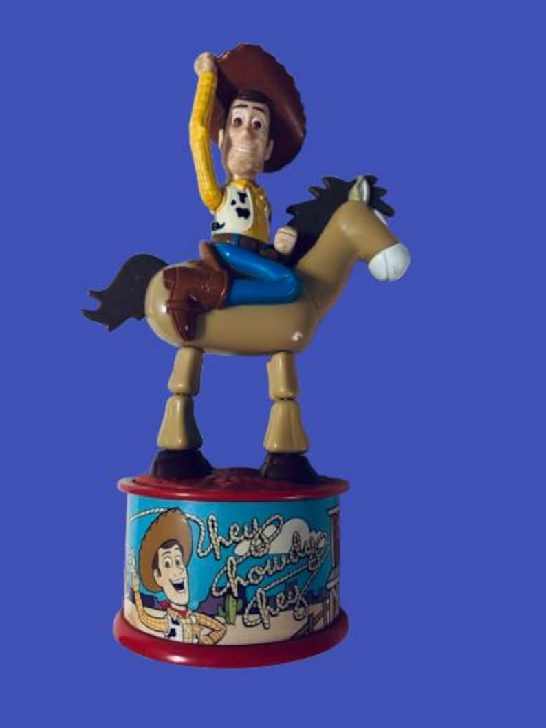 May include: A Woody toy figure riding a horse, with a brown hat and yellow shirt. The base features the text "Hey Howdy Hey" and an image of Woody. The toy is set against a blue background.