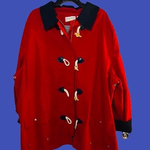 May include: A red duffel coat with a navy blue collar and cuffs, featuring toggle closures and a zipper. The coat is hanging on a black hanger against a blue background. The coat has a classic design.