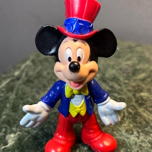 May include: A Mickey Mouse figurine in a patriotic outfit. Mickey wears a red, white, and blue top hat, a blue jacket, red pants, and white gloves. His yellow bow tie adds a pop of color to the classic character.