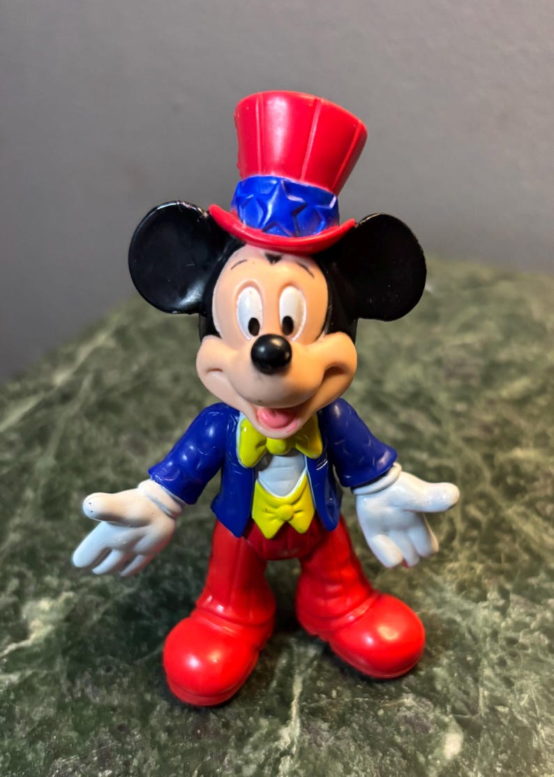 May include: A Mickey Mouse figurine in a patriotic outfit. Mickey wears a red, white, and blue top hat, a blue jacket, red pants, and white gloves. His yellow bow tie adds a pop of color to the classic character.