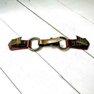 Puede incluir: Una correa de cuero marr&oacute;n con dos clips de metal dorado y un anillo de conexi&oacute;n. La correa est&aacute; dise&ntilde;ada para ser utilizada como un cierre.