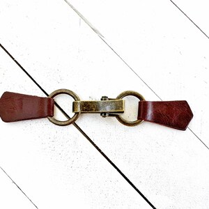Puede incluir: Un cierre de palanca de cuero marr&oacute;n con dos anillos met&aacute;licos y una barra de lat&oacute;n en el centro.