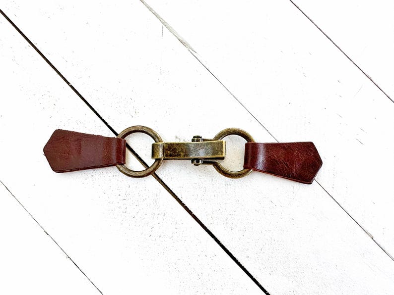 Puede incluir: Un cierre de palanca de cuero marr&oacute;n con dos anillos met&aacute;licos y una barra de lat&oacute;n en el centro.