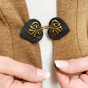 May include: A brown cardigan with a black leather heart-shaped clasp with a gold metal design. The clasp is closed and the cardigan is being held open by two hands.