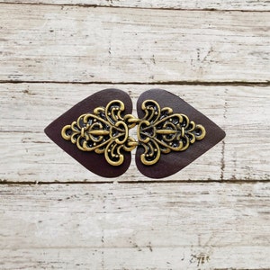 May include: A decorative clasp featuring a dark brown leather base in a heart shape. The clasp has an ornate, antique brass-colored metal design in the center. The clasp is designed to fasten two pieces of fabric or material together.