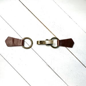 Puede incluir: Una correa de cuero marr&oacute;n con dos anillos met&aacute;licos y un cierre met&aacute;lico. La correa est&aacute; sobre una superficie de madera blanca.