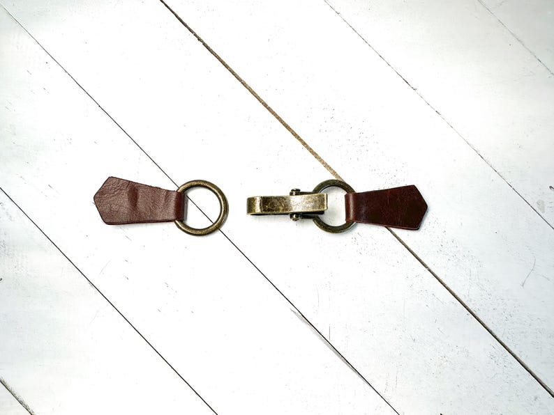Puede incluir: Una correa de cuero marr&oacute;n con dos anillos met&aacute;licos y un cierre met&aacute;lico. La correa est&aacute; sobre una superficie de madera blanca.