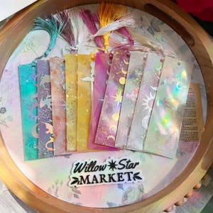 May include: A collection of colorful, handmade bookmarks with tassel accents, featuring celestial and fantasy designs. The bookmarks are displayed on a wooden tray with the text "Willow Star MARKET". Each bookmark has a unique design with glitter and holographic elements.
