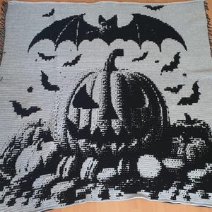 Może przedstawiać: Szary i czarny koc o tematyce Halloween. Koc przedstawia dużą dynię z nietoperzem nad nią i kilkoma mniejszymi nietoperzami. Koc ma frędzelkową krawędź.