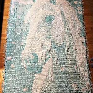 Puede incluir: Una manta tejida con un retrato detallado de la cabeza de un caballo blanco sobre un fondo turquesa. La manta tiene un borde con flecos y un diseño pixelado, creando una apariencia texturizada. El ojo y la melena del caballo están claramente definidos.
