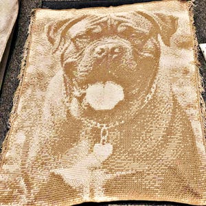 May include: A woven throw blanket featuring a detailed image of a dog's face. The dog has a collar with a heart-shaped pendant. The blanket is primarily tan and cream-colored, with a fringed edge.