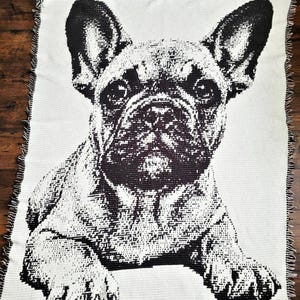 Puede incluir: Una manta tejida de color crema con una imagen en blanco y negro de un bulldog francés. La cara del perro es el punto focal, con sus grandes orejas y sus expresivos ojos. La manta tiene un borde con flecos.